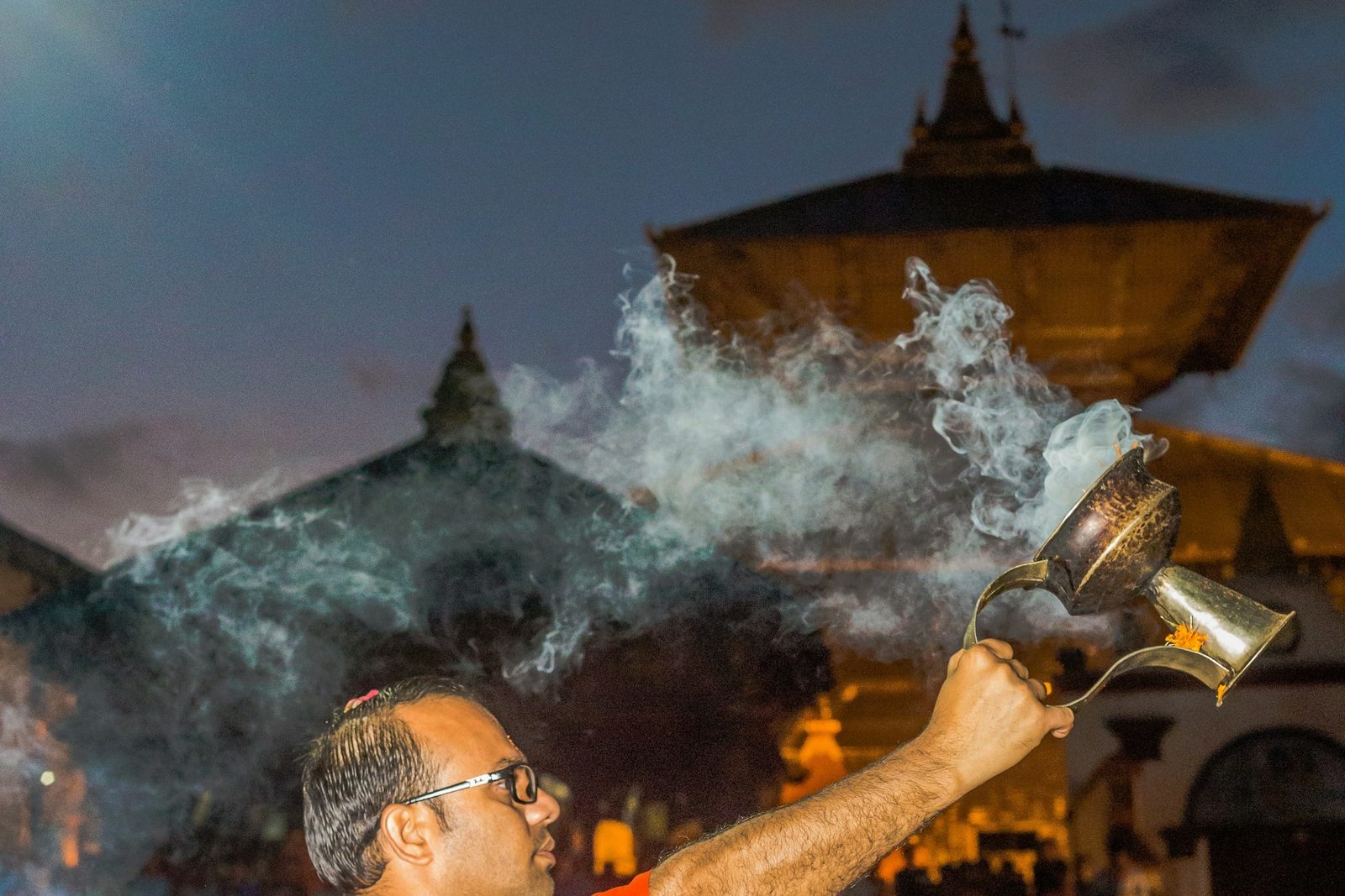 A priest swinging an incense burner during the Pashupatinath aarti, with the temple roofs behind.